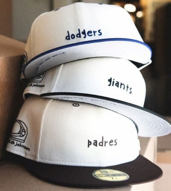 New Era × Aku — three stacked white fitted caps, Dodgers, Giants and Padres, each with a hand-drawn Aku helmet and Micah Johnson's signature